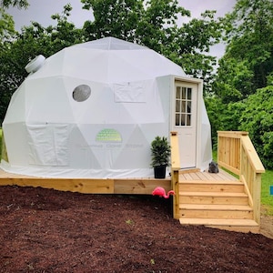 May include: A white geodesic dome with a door, small window, and wooden decking. A small potted plant and a pink flamingo decoration are near the entrance. The dome is set in a grassy area surrounded by trees.