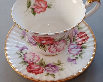 May include: A white porcelain teacup and saucer set featuring a scalloped edge with gold trim. The set is decorated with a floral pattern of red and purple sweet pea flowers and green leaves. The teacup has a curved handle with gold accents.