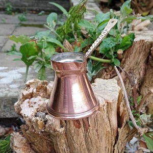 May include: A small copper coffee pot with a long, thin handle. The pot is sitting on a tree stump.