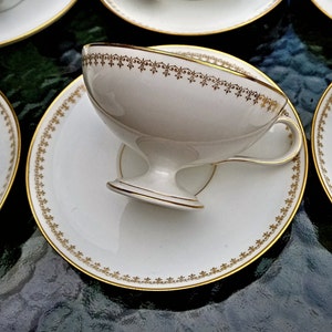 May include: A white porcelain teacup with a gold trim and a delicate floral pattern. The teacup is sitting on a white saucer with a gold rim.