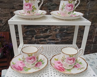 May include: Four teacups and saucers with a floral pattern of pink roses on a white background. The teacups and saucers are displayed on a white metal shelf.