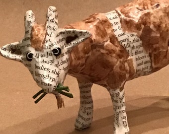 May include: A brown and white paper mache goat sculpture with black eyes. The goat is eating green leaves. The sculpture is made from recycled paper and has a textural surface.