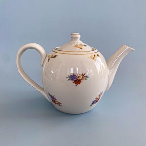 Może przedstawiać: Biały ceramiczny imbryk z zaokrąglonym korpusem, zakrzywionym uchwytem i dzióbkiem. Imbryk posiada kwiatowe wzory w kolorze czerwonym, fioletowym i żółtym. Złote wykończenia zdobią pokrywę i obręcz. Jasnoniebieskie tło.