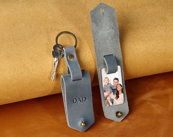 May include: Two gray leather keychains with a photo insert and the word "DAD" embossed on one. The keychain on the left has a silver key ring and two keys attached. The keychain on the right has a photo of a family of three.