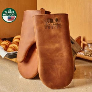 May include: Two brown leather oven mitts with the text "Mom's kitchen" and kitchen utensils. The mitts are made of a smooth, durable material. The image also includes a basket of croissants.