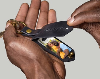 May include: Black leather keychain with a photo insert featuring a father and daughter. The keychain has a flap that opens to reveal the photo and the word "DADDY" is embossed on the flap.