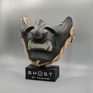 Puede incluir: Una máscara de samurái negra con dientes dorados y una cuerda, exhibida sobre una base negra. La base tiene el texto "GHOST OF TSUSHIMA" en blanco.