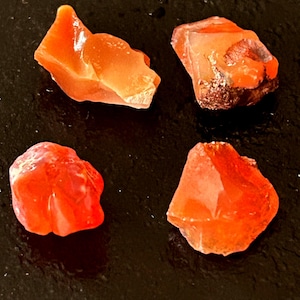 May include: Four raw, unpolished carnelian stones with a vibrant orange-red hue. Each piece displays a translucent, crystalline texture with irregular, jagged edges and natural facets that catch the light, showcasing deep shades of sunset orange.