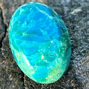 May include: A smooth, oval-shaped turquoise cabochon with a blue and green marbled pattern. The stone has a polished finish and is set against a rough, gray background.