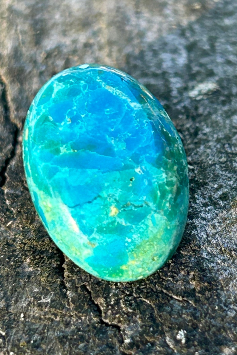 May include: A smooth, oval-shaped turquoise cabochon with a blue and green marbled pattern. The stone has a polished finish and is set against a rough, gray background.