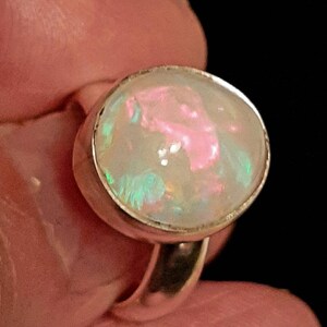 May include: A silver ring with a large oval opal gemstone. The opal has a white base with pink, green, and blue flashes of color.