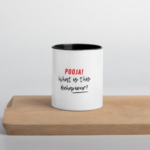 Könnte beinhalten: Eine weiße Keramiktasse mit schwarzem Rand. Die Tasse trägt den Text "POOJA! What is this Behaviour?" in Schwarz und Rot.