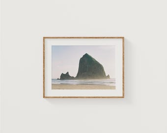 May include: A photograph of a large, green rock formation rising out of the ocean. The rock is surrounded by water and sand, and the sky is a light blue.