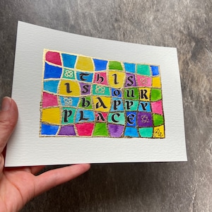 May include: A colorful artwork on textured white paper, featuring the phrase "This is our happy place." The text is in a unique font, set against a mosaic of vibrant squares in blue, yellow, pink, green, and purple, with gold accents.