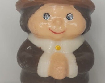 May include: A ceramic pilgrim figurine. It has a brown hat with a yellow buckle, a white collar, and a brown coat. The figurine has a smiling face with blue eyes and a red mouth. The figure is wearing brown trousers and black shoes.