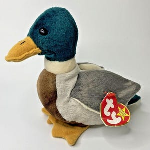 May include: A plush mallard duck toy with a teal head, yellow beak, and brown and gray body. The toy has a white neck and yellow feet. A red tag with white text is attached to the duck.