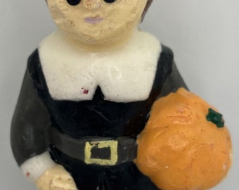 May include: A vintage candle in the shape of a pilgrim figure, with a black hat and outfit, and a white collar. The figure holds an orange pumpkin. This decorative candle is a vintage item.