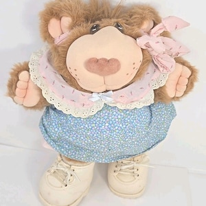 May include: A plush bear doll with brown fur, wearing a blue floral dress, a pink collar with lace trim, and white sneakers. The bear has a large pink nose, small black eyes, and a pink bow on its head. A charming vintage toy.