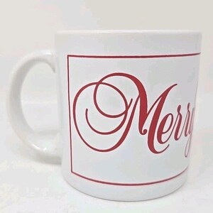 May include: White ceramic mug with a curved handle. The mug features the word "Merry" in red script within a red rectangular border. The design is simple and festive, suitable for holiday use.