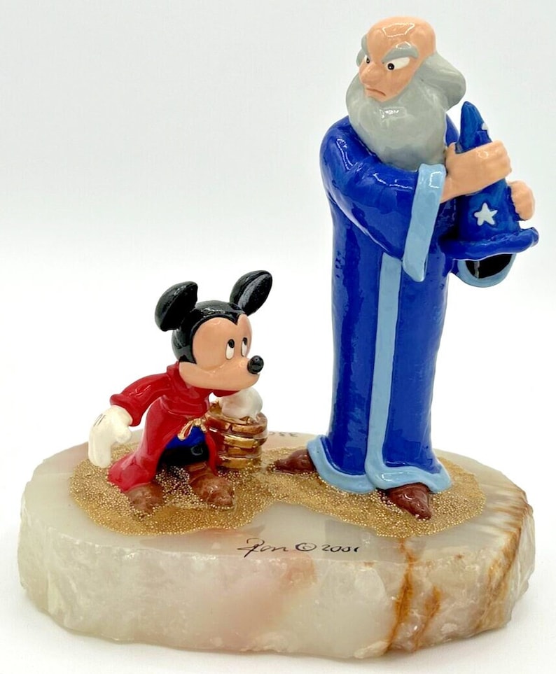 May include: A figurine featuring Mickey Mouse in a red robe and a blue-robed wizard. The base is a light-colored stone with gold glitter accents. The wizard holds a blue hat with a white star. The figurine is signed "Jon &copy; 2001".