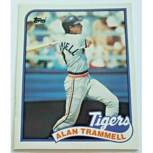 May include: A Topps baseball card featuring Alan Trammell of the Detroit Tigers in a batting stance. The card has a blue border and the Tigers logo. The player is wearing a gray and white uniform with orange and black accents.