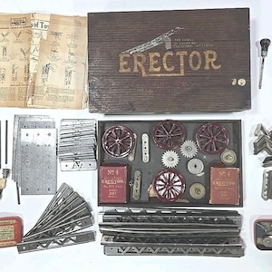 May include: A vintage Erector Set with various metal parts, including wheels, gears, and beams. The set includes a wooden box with the word "Erector" in gold lettering, instruction manuals, and small red boxes.