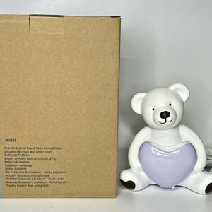 May include: White ceramic bear diffuser with a purple heart. The diffuser is plugged in and has a cord. The box for the diffuser is brown and has the text "PartyLite" and "P91333" on it.