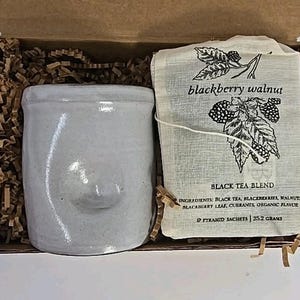 May include: A gift set in a cardboard box containing a light gray ceramic mug and a tea bag. The tea bag is labeled "blackberry walnut" and contains a black tea blend. The box is filled with brown paper shreds.