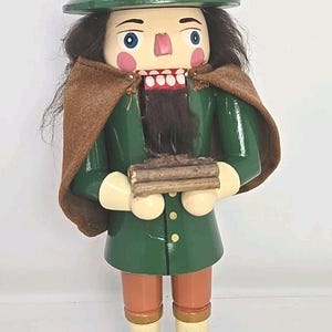 May include: A wooden nutcracker figurine in a green coat and hat, holding a bundle of wood. The figurine has a long black beard, rosy cheeks, and a wide open mouth with white teeth. The hat has a gray band.