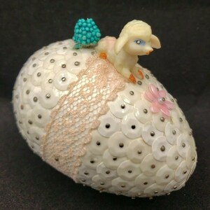 May include: A white Easter egg covered in sequins with a pink and white lace ribbon and a small white lamb figurine on top. The lamb has blue eyes and a pink nose.