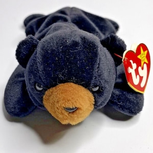 May include: A black plush bear toy with a brown snout and small, round eyes. The bear has a red and white tag attached to its ear. The toy is lying down on a white surface.
