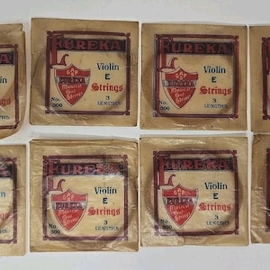 May include: Eight vintage packages of Eureka Violin E Strings. Each package has a brown and red label with the text "Eureka" and "Violin E Strings". The packages are sealed in clear cellophane.