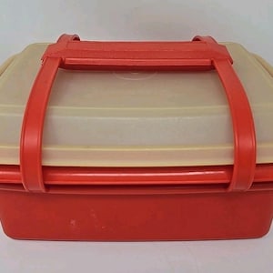 May include: A red and beige rectangular food storage container with a matching lid and a red handle. The container is made of plastic and appears to be vintage. The lid has a slightly translucent appearance.