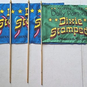 May include: A collection of small flags on wooden poles. Three blue flags feature the word "Stampede" in yellow and red, with stars and a ribbon design. A green flag displays "Dolly Parton's Dixie Stampede Dinner & Show."