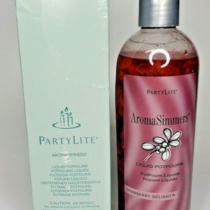 May include: A PartyLite Aroma Simmers liquid potpourri bottle in Cranberry Delight. The bottle is clear with a red liquid and a silver lid. The label is pink with white text and a cranberry graphic.