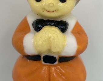 May include: A ceramic figurine of a pilgrim boy. The boy wears a black hat with an orange band, an orange jacket with white trim, a black bow tie, and black shoes. The figurine is a Thanksgiving decoration.