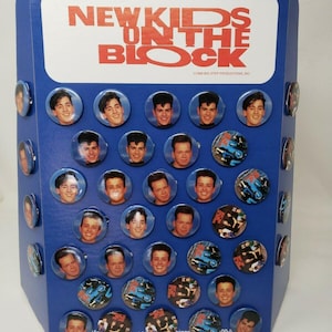 May include: A blue display stand featuring the text "NEW KIDS ON THE BLOCK" in red. The stand holds numerous circular buttons with images of the band members and graphics. The buttons are attached to the stand.