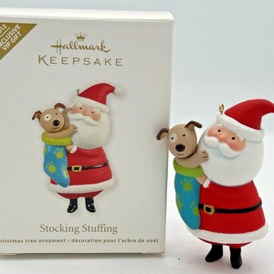 May include: A Christmas tree ornament by Hallmark, depicting Santa Claus embracing a brown dog in a blue stocking. The ornament is red, white, and black with gold details. The box reads "Stocking Stuffing" and "2012 Exclusive VIP Gift."