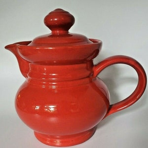 May include: A red ceramic teapot with a lid and a handle. The teapot has a rounded body and a slightly flared spout.