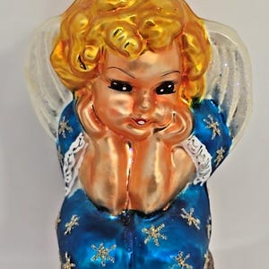 May include: A vintage Christmas ornament shaped like a cherubic angel. The angel has golden hair, a blue dress with gold star accents, and white wings. The ornament has a gold metal cap and a loop for hanging.