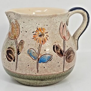 May include: A ceramic pitcher with a cream-colored speckled finish. The pitcher is adorned with hand-painted flowers in orange, pink, and blue. The handle is dark blue, and the base has a green stripe.