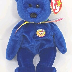 May include: A plush blue teddy bear with a black nose and eyes. It has a colorful ribbon around its neck and a circular "Official Club" patch. A red heart-shaped tag is attached.