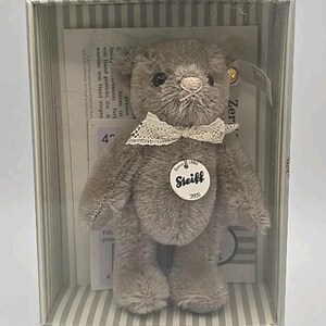 May include: A gray Steiff teddy bear in a clear box with a striped background. The bear has a lace collar and a circular tag that reads "Steiff 2020." The box contains a printed paper with text. The bear is a collectible item.