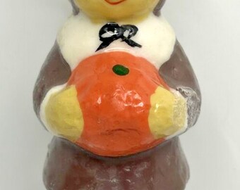 May include: A vintage candle in the shape of a pilgrim figure, holding an orange pumpkin. The candle has a brown hat, coat, and trousers, with black shoes. The figure has a yellow face and a black bow tie. A festive Thanksgiving decoration.