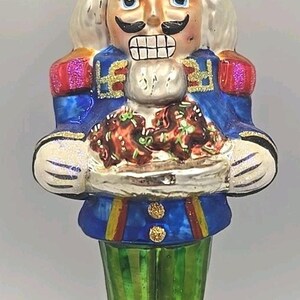May include: A detailed glass Christmas ornament shaped like a nutcracker soldier. The ornament features a blue jacket, green striped pants, and a black hat with gold accents. It holds a plate of food.