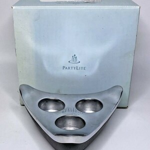 May include: Silver candle holder with three candle wells in a triangular shape. The holder has a wood base and the text "PartyLite" is printed on the box.