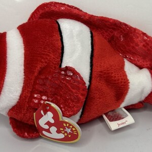 May include: A plush toy clownfish with red, white, and black stripes. The fish has large blue eyes and a small blue mouth. The toy has a tag that says "Beanie Baby" and "Jester".