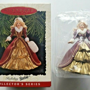 May include: Holiday Barbie Collector's Series ornament. The doll is wearing a burgundy and gold gown with white fur trim. The ornament is in its original packaging, with the words "Holiday Barbie" and "Collector's Series" visible.