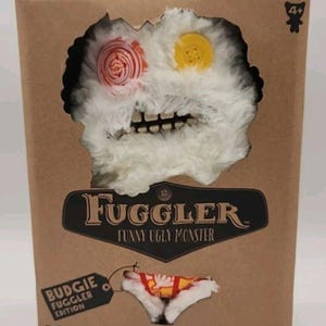 May include: A brown cardboard box featuring a Fuggler plush toy. The toy has a white, shaggy body, a toothy grin, and button eyes. The box reads "Fuggler Funny Ugly Monster" and "Budgie Fuggler Edition."