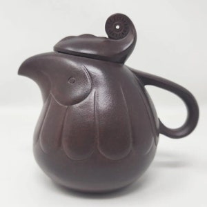 May include: A dark brown ceramic teapot with a unique, organic design. The teapot features a curved spout, a rounded body, and a handle. The lid has a decorative swirl. The overall shape resembles a flower or a bird.